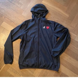 vindjacka Comme des Garçons x K-Way - Mörkblå  vindjacka från Comme des Garçons Play i samarbete med K-Way. Jackan har huva, hel dragkedja och elastiska muddar. Ikoniskt rött hjärta på bröstet och ryggen samt K-Way-logga framtill. Perfekt för dig som gillar streetwear och stilrena detaljer.