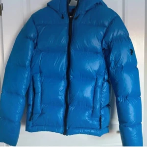 Blå Peak Performance dunjacka S - Säljer en blå dunjacka från Peak Performance i storlek S. Jackan har en puffig look, huva och dragkedja framtill. Perfekt för kalla dagar och har en riktigt snygg streetstil. Klassisk logga på ärmen och två fickor med dragkedja.