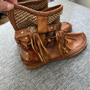 Karma of Charme bruna boots med fransar - Unika boots från Karma of Charme i brunt skinn med handgjorda detaljer, fransar och dekorativa metalldetaljer. Ovandelen har flätad textil och coola stjärnprydnader. Perfekta för dig som vill sticka ut med boho-vibe och italiensk design. Inköpta i Ibiza. Använda endast 2 gånger. I väldigt gott skick och inget slitage. Nypris då 300 euro.