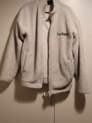 Vit teddyjacka Los Angeles S - Mysig vit teddyjacka i storlek S med broderad Los Angeles-text fram och stort tryck på ryggen. Jackan har dragkedja, höga fickor och en fluffig, varm känsla. Perfekt för dig som gillar streetstyle och vill ha något som sticker ut.