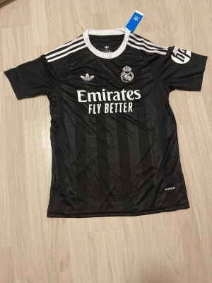 Svart Real Madrid Adidas tröja - Säljer en svart Real Madrid tröja från Adidas med vita detaljer och logga på bröstet. Tröjan har klassiska tre ränder på axlarna, rund halsringning och tryck med 'Emirates Fly Better' på framsidan. Tillverkad i polyester och har AEROREADY-teknologi.