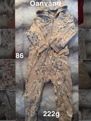 Beige blommig byxdress från Newbie - Supersöt beige byxdress från Newbie med huva och dragkedja framtill. Täckande blommönster i vitt och grönt, ribbade muddar vid ärmar och benslut. Perfekt för chill dagar hemma eller när du vill vara extra comfy.🌸 Oanvänd. Endast upptvättad. Storlek 86