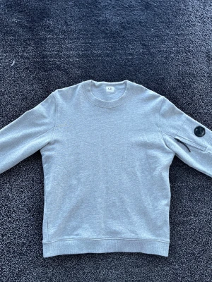 C.P. Company sweatshirt - Säljer en stilren grå sweatshirt från C.P. Company. Tröjan har rund hals, lång ärm och en ikonisk detalj med svart lins och dragkedja på ena ärmen. Perfekt för dig som gillar minimalistisk design med en twist. Storlek M, skick 10/10 som ny, äkta. Pris går att diskutera✅