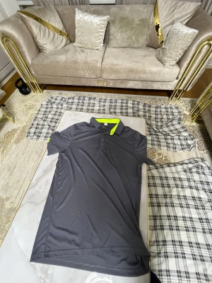 Mörkgrå pikétröja med neongul krage - Snygg mörkgrå pikétröja med korta ärmar och knappar framtill. Kragen har en cool neongul insida som ger en sportig touch. Tillverkad i ett lätt och mjukt syntetmaterial som andas bra. Perfekt för dig som gillar stilrena plagg med en twist.