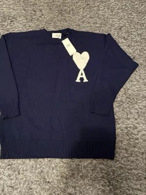 Mörkblå stickad tröja från Ami - Säljer en mörkblå stickad tröja från Ami med ikoniskt vitt hjärta och A på bröstet. Tröjan har rund hals, ribbade muddar och är långärmad. Perfekt för dig som vill ha en clean men ändå statement look. Priset kan diskuteras 