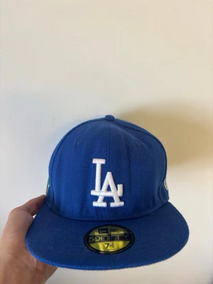 Blå LA-keps från New Era 59FIFTY - Säljer en blå New Era 59FIFTY keps med LA-logga framtill och broderad text 'Greetings from Los Angeles' samt palmer på sidan. Klassisk rak skärm och snygga detaljer i vitt, grönt och gult. Perfekt för dig som gillar streetwear och vill sticka ut.