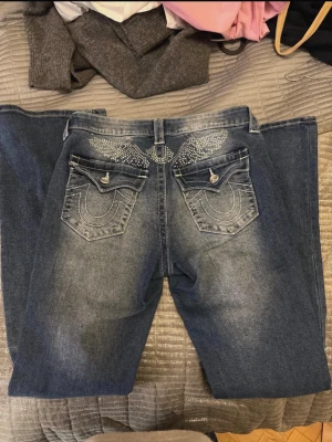 Blå jeans med stenar och broderi - Säljer ett par blå jeans från True Religion med snygga rhinstone vingar på baksidan!  och coolt broderi! Klassisk femficksmodell med raka ben och kontrastsömmar. Perfekta för dig som vill sticka ut lite extra. Originalt köpt för 2500sek! 