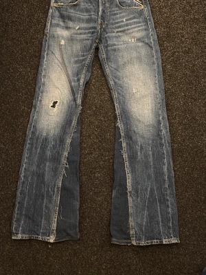 Custom flare replay jeans - Säljer ett par blå jeans med bootcut passform och slitna detaljer. Jeansen har hål och slitningar på benen för en riktigt avslappnad vibe. Klassisk femficksmodell med kontrastsömmar och ljusare tvätt på framsidan. Perfekta för dig som gillar en vintage look.