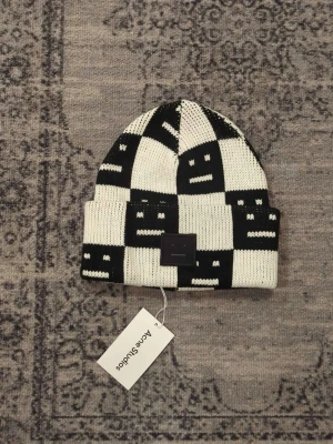 Rutig svartvit mössa Acne Studios - Säljer en stickad mössa från Acne Studios i svartvitt rutigt mönster med deras ikoniska ansikte. Mössan är i ullblandning och har en ribbad kant. Perfekt accessoar för att lyfta din look.