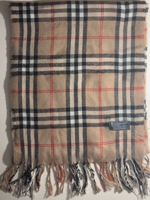 Rutig beige halsduk från Burberrys - Klassisk halsduk från Burberrys i 100% kashmir. Beige botten med ikoniskt rutigt mönster i svart, vitt och rött. Mjukt material och fransar i ändarna. Perfekt accessoar för att lyfta din outfit med en tidlös brittisk vibe. 