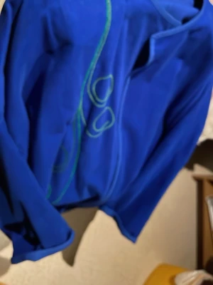 Blå omlott kappa från Unica Fashion - Säljer en blå kappa från Unica Fashion med omlott-design och coola gröna broderade detaljer på framsidan. Jackan har lång ärm och stora fickor framtill. Perfekt för dig som vill sticka ut med färg och unik stil. Säljs på fler sidor. Känns som sammet fast det är bomull