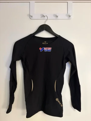 Dam svart långärmad träningströja  - Svart långärmad träningströja från InterSport i storlek M (passar S) Tröjan har Intersport-logga på bröstet, guldfärgade detaljer och mönster på ryggen samt diskreta loggor på ärmarna. Tillverkad i syntetmaterial som andas, perfekt för sport och träning. 