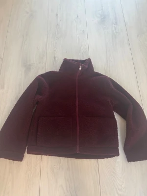 Vinröd teddyjacka med dragkedja - Mysig vinröd teddyjacka med hög krage och hel dragkedja framtill. Jackan har två stora fickor på framsidan och är tillverkad i ett fluffigt teddy-material som håller dig varm. Perfekt för kyliga dagar och enkel att styla till jeans eller leggings.