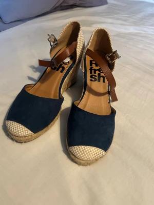 Blå kilklack sandaler med flätad detalj - Säljer ett par snygga sandaler med blå mockaliknande tå, beige flätad häl och kilklack i rep. Skorna har bruna remmar med spänne och öppen tå. Perfekta för sommarens alla tillfällen!