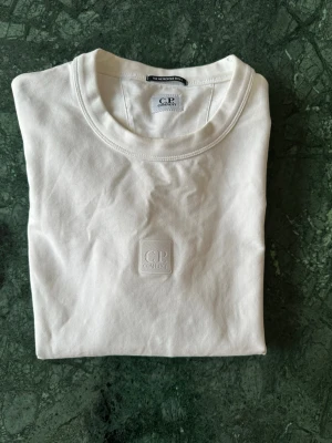 Vit sweatshirt från C.P. Company - Säljer en clean vit sweatshirt från C.P. Company med diskret logopatch på bröstet. Klassisk rund halsringning och långa ärmar. Tillverkad i mjuk bomull som känns skön mot huden. Perfekt för dig som gillar minimalistisk stil. Hör av dig vid funderingar!🤩