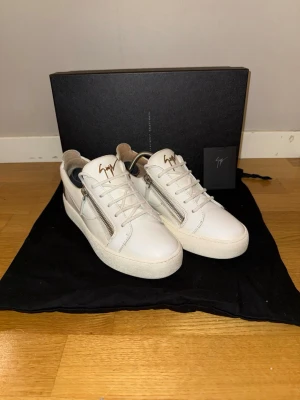 Vita sneakers från Giuseppe Zanotti - Snygga vita sneakers från Giuseppe Zanotti med guldfärgad logga på plösen, dragkedjor på sidorna och svarta lackdetaljer på hälen. Skorna har snörning och är tillverkade i skinn med en chunky vit sula. Perfekta för dig som vill sticka ut med stilrena detaljer. Storlek 40 men passar 42