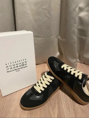 Maison Margiela sneakers svart - Maison Margiela  sneakers i svart med kontrasterande beige snörning och gummisula. Skorna har en klassisk rund tå och är tillverkade i en mix av skinn och mocka. Snygg retrostil med diskreta detaljer och vit insida. Perfekt för dig som gillar stilrena och tidlösa sneakers.