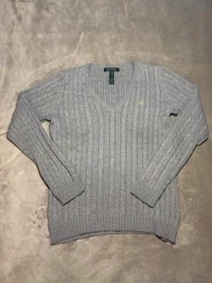 Grå kabelstickad tröja från Ralph Lauren - Kabelstickad grå tröja från Lauren Ralph Lauren med v-ringning och diskret RL-broderi i gult på bröstet. Tröjan har lång ärm, ribbade muddar och är gjord i mjuk bomullsblandning. Perfekt för lager-på-lager under kyliga dagar.