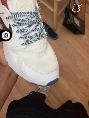 Nike vita sneakers med svart swoosh - Säljer ett par vita Nike sneakers med svart swoosh på sidan, röda och limegula detaljer på hälen samt blått framtill på sulan. Skorna har randiga snören och är tillverkade i syntetmaterial och skinn. Perfekta för dig som gillar klassisk streetwear.