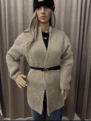 Beige fluffig cardigan jacka med bälte - Mysig och fluffig beige cardigan jacka med lång ärm och öppen front. Jackan har en rak passform och är markerad i midjan med ett svart bälte med gulddetalj. Perfekt för dig som gillar en chill och trendig look.
