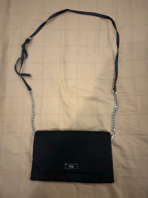 Svart kuvertväska från Fiorelli - Stilren svart kuvertväska från Fiorelli med kedjerem och detaljer i skinnimitation. Väskan har magnetlås, innerfack med dragkedja och en praktisk handledsrem på baksidan. Perfekt storlek för det viktigaste och enkel att bära crossbody.