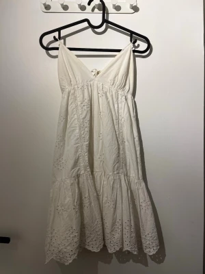 Vit kort klänning med broderi - Söt vit kort klänning med tunna axelband och v-ringning. Klänningen har broderade blommönster och volangkant nertill. Tillverkad i bomull och har knytband i ryggen för extra detalj. Perfekt för sommarens alla tillfällen.