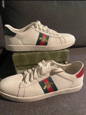 Kvitto finns. Gucci Ace sneakers med bi-detalj - Säljer ett par vita Gucci Ace sneakers i skinn med klassiska gröna och röda ränder på sidan samt en guldfärgad bi-detalj. Baksidan har metallicrött och metallicgrönt parti. Snygg snörning och platt sula med Gucci-logga undertill.