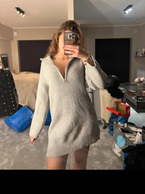 Grå stickad klänning med half zip - Mysig grå stickad tröja med lång ärm och half zip-dragkedja framtill. Tröjan har en loose passform och är perfekt för kyliga dagar. Ribbstickad struktur och hög krage som kan vikas ner.