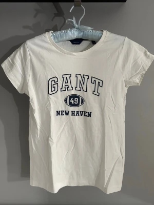 Vit GANT t-shirt med tryck XS - Vit t-shirt från GANT i storlek XS med klassiskt tryck framtill där det står 'GANT 49 New Haven'. T-shirten har rund halsringning och korta ärmar. Tillverkad i mjuk bomull för en skön känsla och enkel stil.