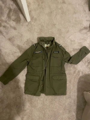 Olivgrön parkajacka Polo Ralph Lauren - Säljer en olivgrön parkajacka från Polo Ralph Lauren Denim & Supply. Jackan har flera fickor framtill, coola patchar och detaljer på axlarna samt hög krage. Tillverkad i bomull och har klassisk militärstil. Perfekt för dig som gillar utility-vibe. Storlek M (barn)