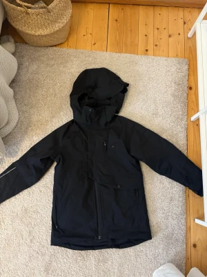 Svart skidjacka från Everest  - Säljer en svart skidjacka från everest med huva, dragkedjor och flera fickor framtill. Jackan har justerbara ärmslut med kardborre och reflekterande detalj på ryggen. Perfekt för skidåkning och vintern. Snö lås i midja och ärmar, huva som passar över hjälm och avtagbar, innerficka, ficka för liftkort på vänster ärm, många fickor, vadderad, vatten och vind tät. Storlek 140. Nypris ca 1500.