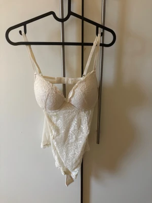 Vit spetsbody med knäppning - Säljer en vit body med tunna axelband och vadderade kupor. Bodyn är täckt av blommig spets och har praktisk knäppning i grenen. Perfekt för att styla under en kavaj eller till jeans för en trendig look. Den är lite knopprig vid bysten men det går att fixa.