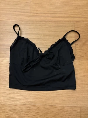 Svart linnetopp med spets från SHEIN, storlek XS - Svart croppad linnetopp från SHEIN i storlek XS med tunna axelband, spetskant upptill och en söt rosett framtill. Toppen är i mjukt material och har en enkel, stilren look som passar perfekt till jeans eller kjol.