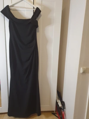 Svart offshoulder långklänning - Säljer en elegant svart långklänning med offshoulder-ringning och figurnära passform. Klänningen har en snygg drapering framtill och är tillverkad i ett mjukt, följsamt material som ger en lyxig känsla. Perfekt för dig som vill sticka ut på bal eller fest.