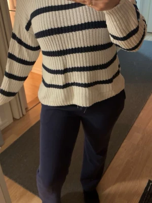 Randig stickad tröja i vitt och blått - Säljer en mysig stickad tröja med breda marinblå ränder på vit botten. Tröjan har långärmade ärmar och en ribbad struktur som ger en skön känsla. Perfekt för dig som gillar klassisk och chill stil.