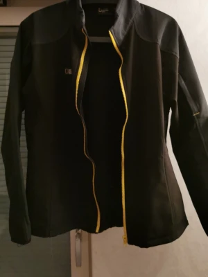 Svart vindjacka från National Geographic - Svart vindjacka från National Geographic med gul dragkedja och diskret logga på bröstet. Jackan har lång ärm, hög krage och är tillverkad i ett slätt syntetmaterial. Snygg och enkel design som passar perfekt till vardagsoutfiten.