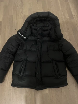 Svart pufferjacka från Gabos London - Säljer en svart pufferjacka från Gabos London med hög krage och huva. Jackan har stora, quiltade partier, dragkedja och knappar framtill samt praktiska fickor med detaljer. Perfekt för kalla dagar och riktigt snygg streetstil.