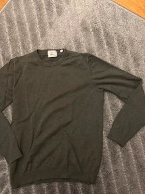 Mörkgrå tröja från Bläck - Säljer en stilren mörkgrå tröja från Bläck i klassisk modell med rund hals och långa ärmar. Passar perfekt för dig som gillar enkel och clean stil. Ribbstickade muddar vid hals, ärmslut och nederkant.