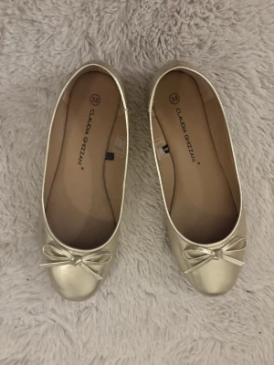 Guldiga ballerinaskor med rosett - Snygga ballerinaskor i guld från Claudia Ghizzani. Skorna har rund tå och en söt rosettdetalj framtill. Perfekta för dig som vill ha en stilren och glansig look. Tillverkade i syntetmaterial och passar till många olika outfits.
