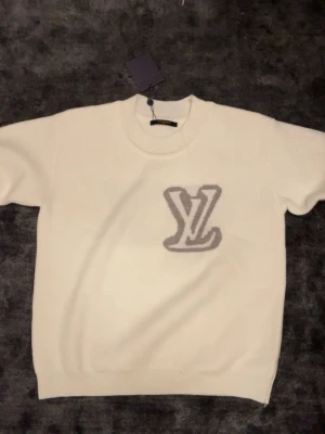 Vit stickad tröja från Louis Vuitton - Exklusiv vit stickad tröja från Louis Vuitton med rund halsringning och ribbade muddar. Tröjan har en mjuk känsla och subtilt mönster i grått framtill. Perfekt för dig som vill ha en clean och lyxig look.