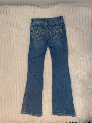 Blå bootcut jeans med broderade fickor - Säljer ett par blå bootcut jeans med snygga broderade detaljer och paljetter på bakfickorna. Jeansen har normal passform, klassisk femficksdesign och är tillverkade i mjukt denimtyg. Perfekta för dig som vill ha ett par jeans med lite extra detaljer.