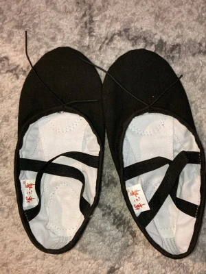 Svarta balett skor med resårband - Svarta balett skor i tyg med rund tå och vita innersulor. Skorna har elastiska korsade band över foten för extra stöd och en platt sula med förstärkning under tå och häl. Perfekta för dans eller balettträning. Dom är aldrig använda.