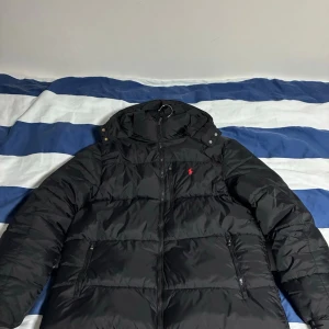 Svart pufferjacka från Polo Ralph Lauren - Svart pufferjacka från Polo Ralph Lauren med huva och dragkedja framtill. Jackan har två sidofickor med dragkedja och ett rött Polo-logo broderat på bröstet. Perfekt för kalla dagar och har en schysst, lite oversized look. Sparsamt använd är storlek XL men passar även L