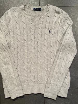 Ljusgrå kabelstickad tröja Polo Ralph Lauren - Säljer en ljusgrå kabelstickad tröja från Polo Ralph Lauren i storlek S. Tröjan har rund hals, långärmade ribbade muddar och den klassiska broderade loggan på bröstet. Mjuk och skön stickad bomull, perfekt för kyliga dagar.