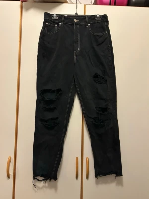Svarta ripped jeans från H&M Divided - Svarta jeans från H&M Divided med grova slitningar och trasiga detaljer på benen. Kontrastsömmar i beige och klassisk femficksdesign. Rå kant vid benslut för extra edgy vibe. Perfekt för dig som gillar streetstyle och vill sticka ut.