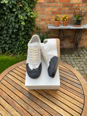 Maison Margiela sneakers med färgstänk - Maison Margiela sneakers i vitt och beige skinn med navyblå färgstänk och detaljer i mocka. Snygg kontrast mellan den ljusa ovandelen och den svarta tån, samt beige gummisula. Klassisk siluett med snörning och rund tå. Sänkt pris vid snabb affär.