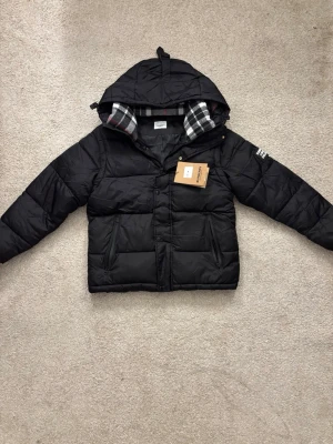 Svart pufferjacka från Burberry - Säljer en svart pufferjacka från Burberry med huva och klassiskt rutigt foder i svart, vitt och lite rött. Jackan har dragkedja och knappar framtill samt två snedställda fickor. Perfekt för kalla dagar och riktigt snygg till streetwear-stilen.