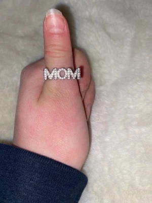 Silverring med stenar och MOM-text - Säljer en snygg silverfärgad ring med glittrande stenar som bildar ordet MOM framtill. På sidorna finns utskurna hjärtan som ger en extra gullig touch. Perfekt som present till någon speciell! Ringen har en modern och stilren design.