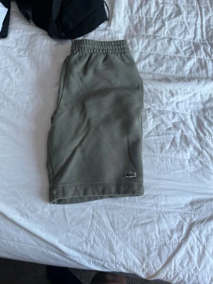 Gröna shorts från Lacoste - Säljer ett par gröna shorts från Lacoste med elastisk midja och den klassiska krokodilloggan broderad längst ner. Sköna och stilrena, perfekta för sommaren. Materialet känns mjukt och andas bra.