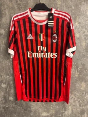 AC Milan Ibrahimovic fotbollströja - Ny XL - Säljer en officiell AC Milan matchtröja från Adidas med Ibrahimovic och nummer 11 på ryggen. Tröjan har klassiska röda och svarta ränder, broderat klubbmärke med stjärna, Fly Emirates-tryck och vita detaljer vid krage och ärmslut. Perfekt för fotbollsfans!